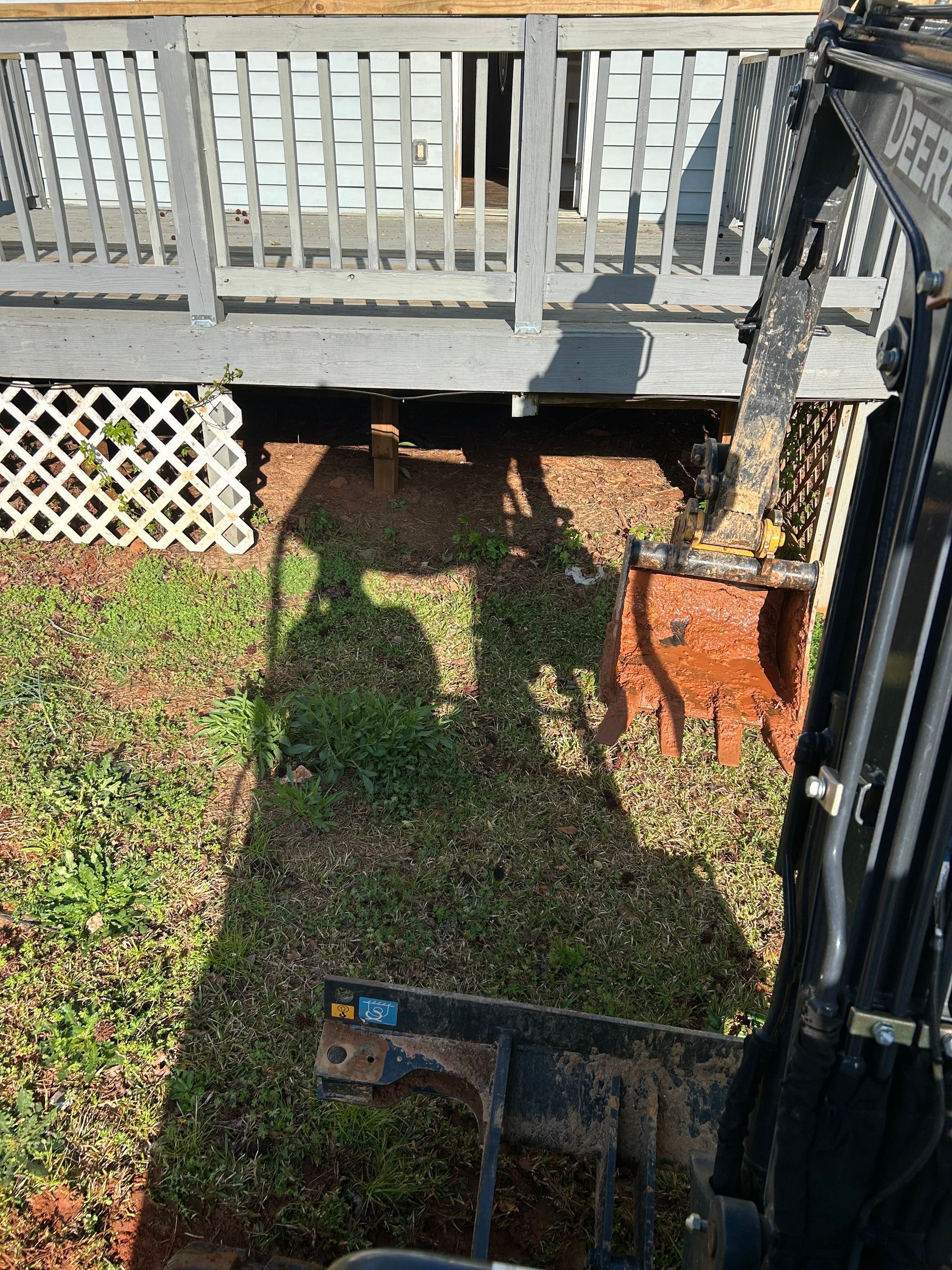 A small excavator digs under a deck, removing dirt. Grass and lattice details are visible in the yard.