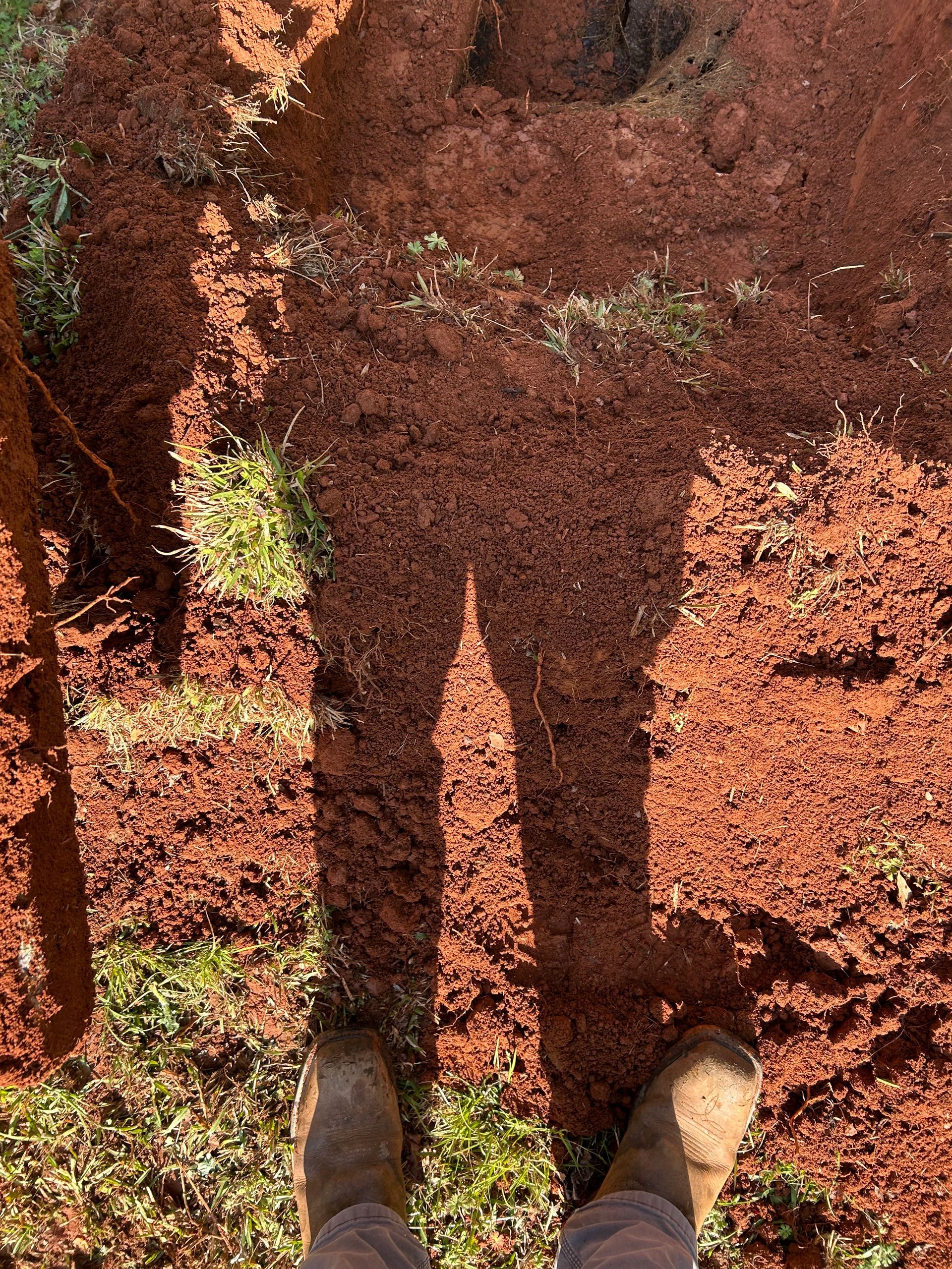 Shadow of person standing in red dirt; their boots visible. Grass patches dot the ground.