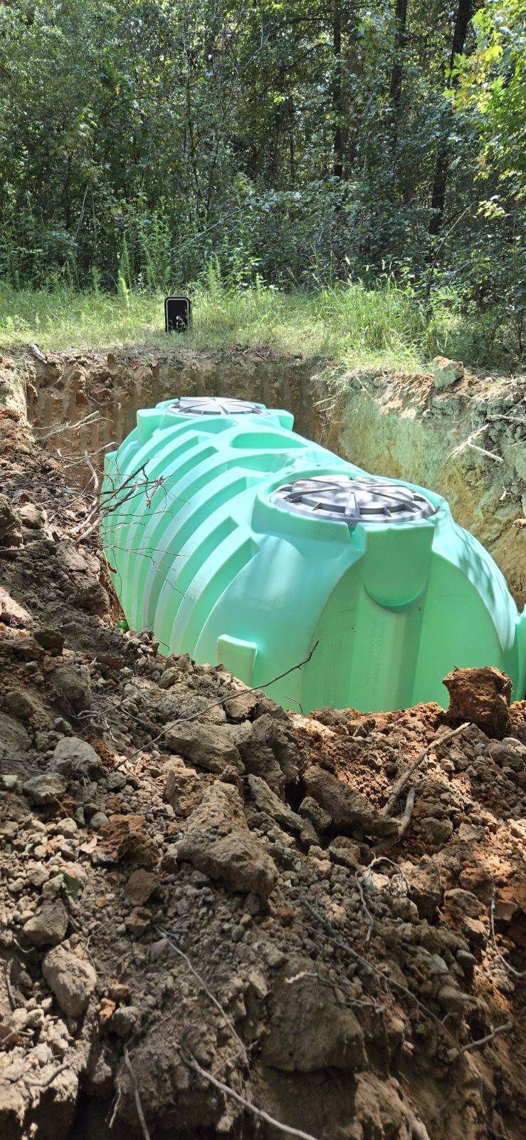 A large, green septic tank sits in a trench dug in the earth. The setting is outdoors with trees in the background.