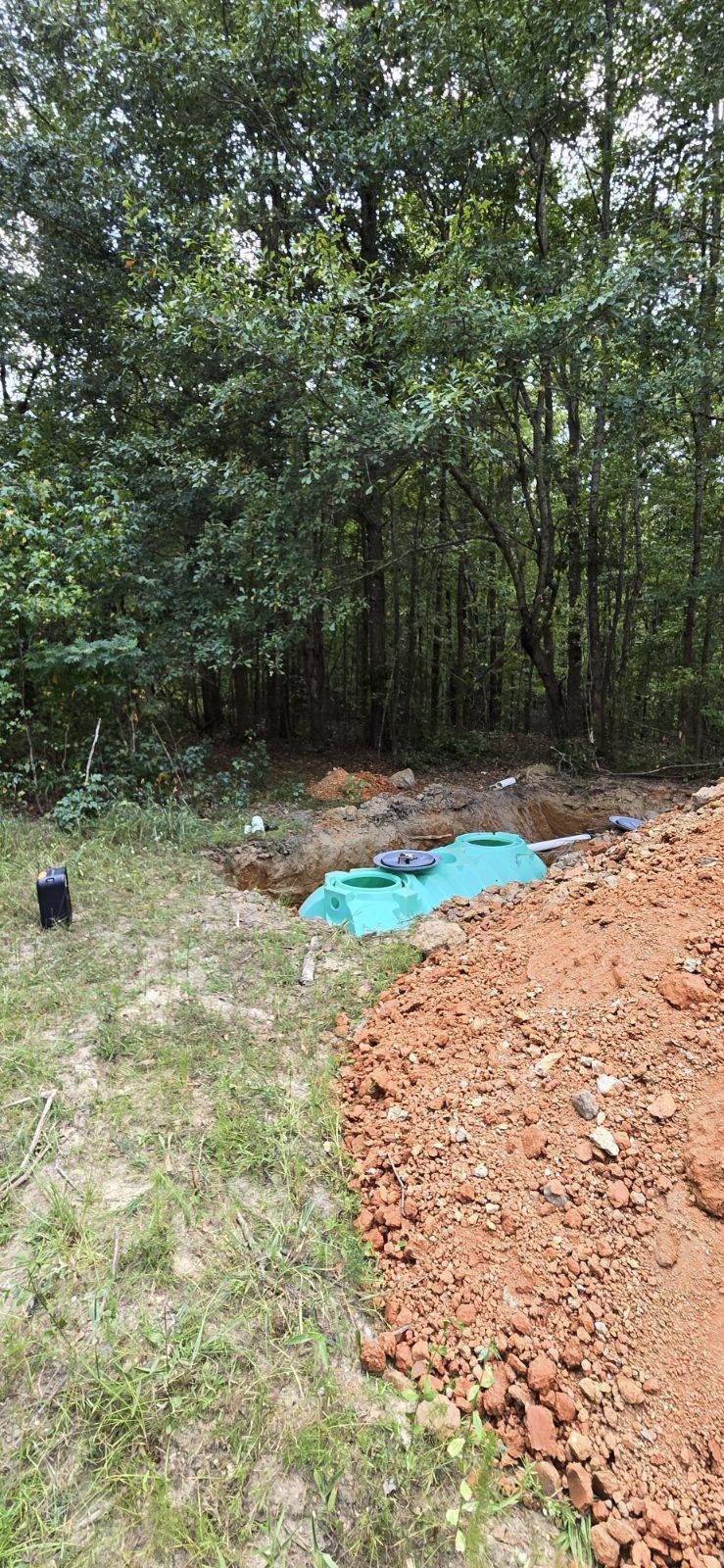 A septic tank installation in a wooded area. A partially buried teal-colored septic tank is surrounded by dirt and grass.