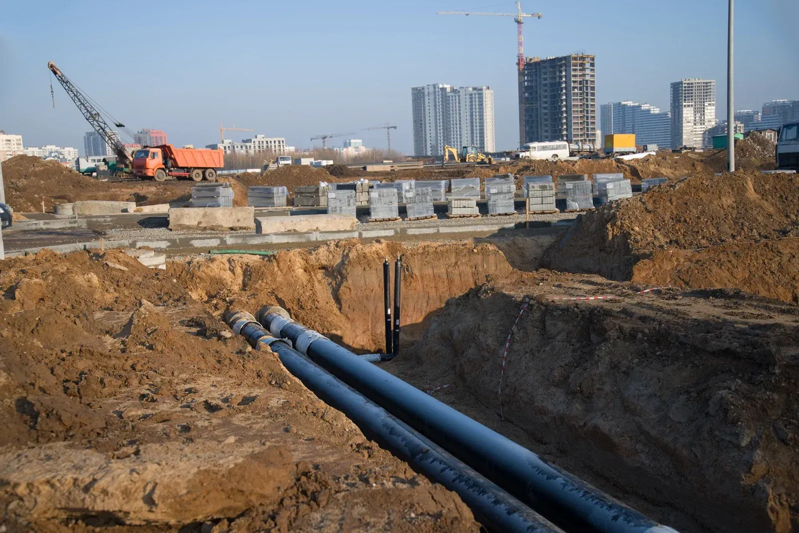 Construction site: buried pipes in a trench, crane, blocks, buildings in the background.