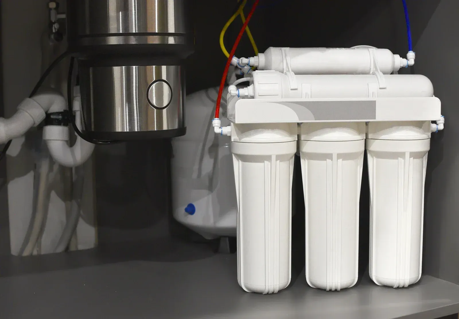 Under-sink water filter system with three white filter cartridges, a garbage disposal, and plumbing.