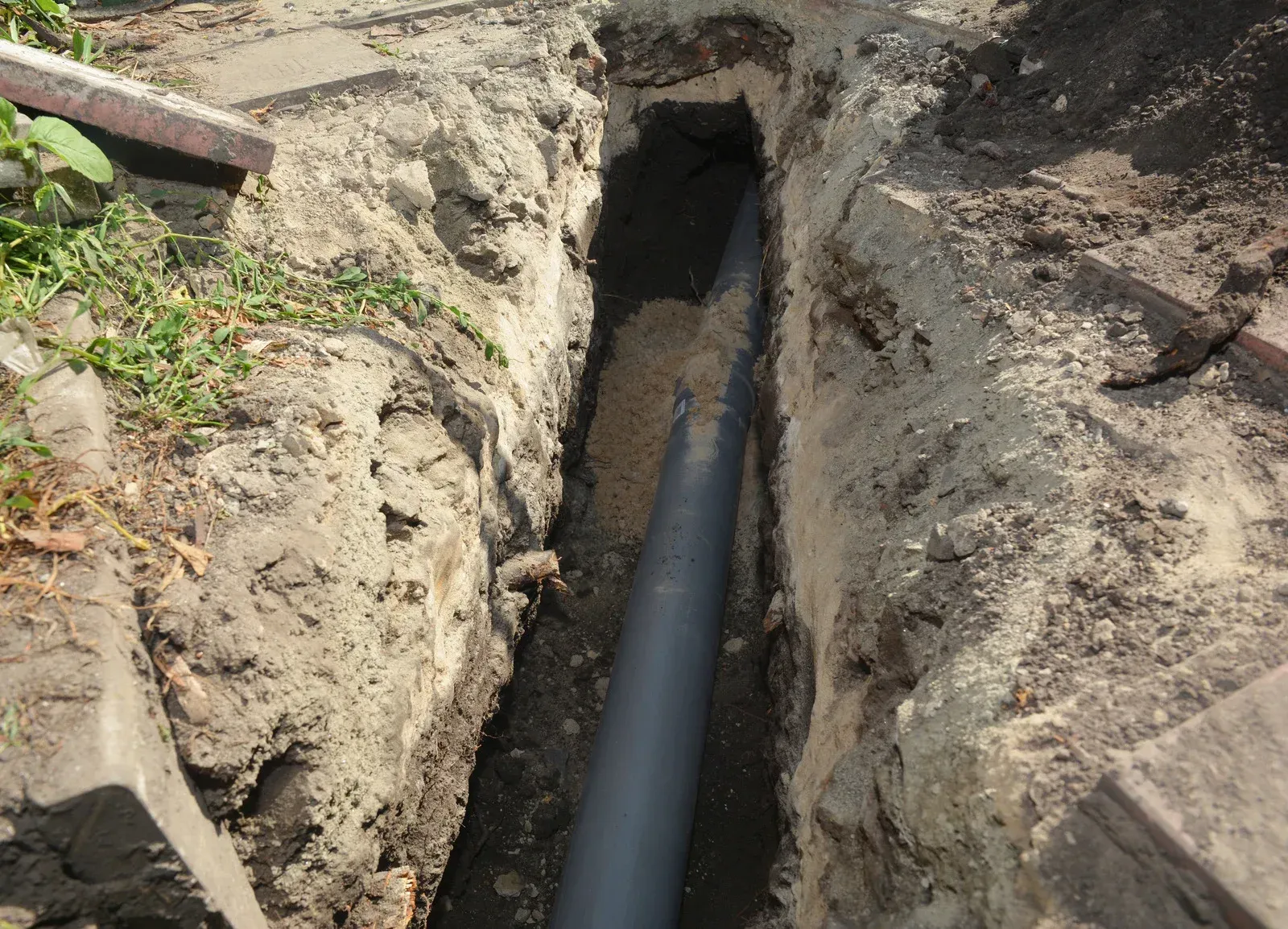 A trench dug in soil containing a dark pipe. The trench is in a yard setting.