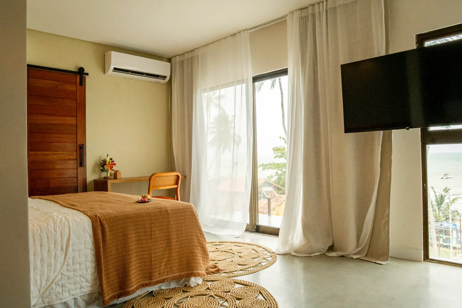 Quarto iluminado com cama, ar-condicionado de parede, cortinas, portas para varanda e TV fixada na parede.