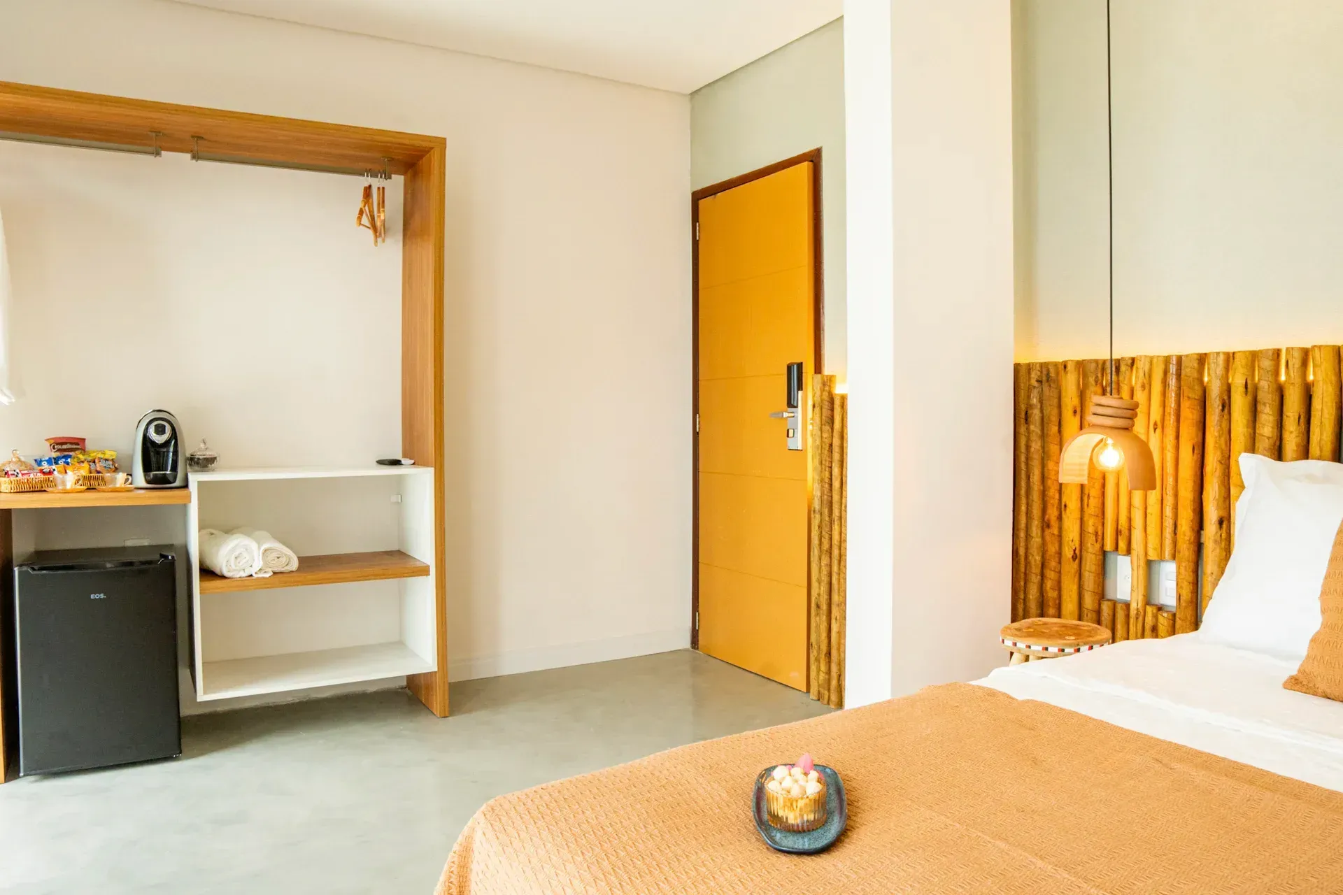 Quarto de hotel moderno com cama, cabeceira de madeira, prateleiras abertas e pequena cozinha.