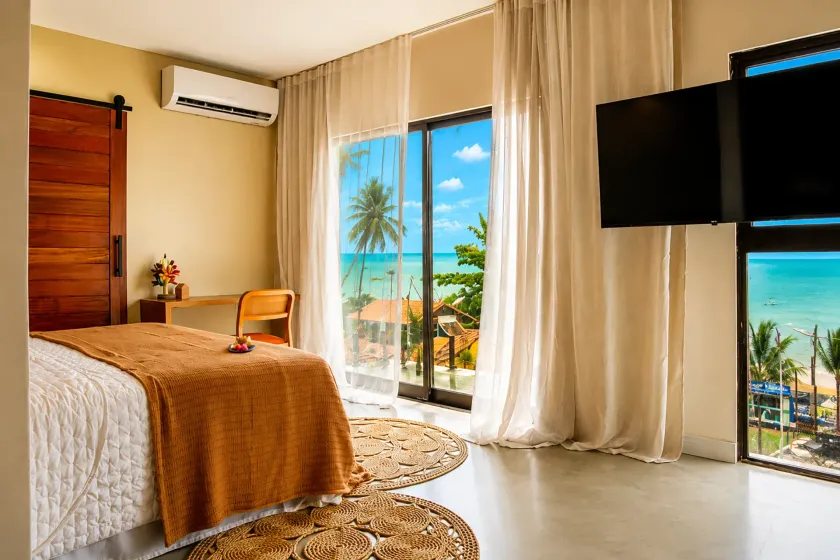 Quarto luminoso com cama, TV de parede e portas de correr que se abrem para uma vista ensolarada de uma praia tropical.