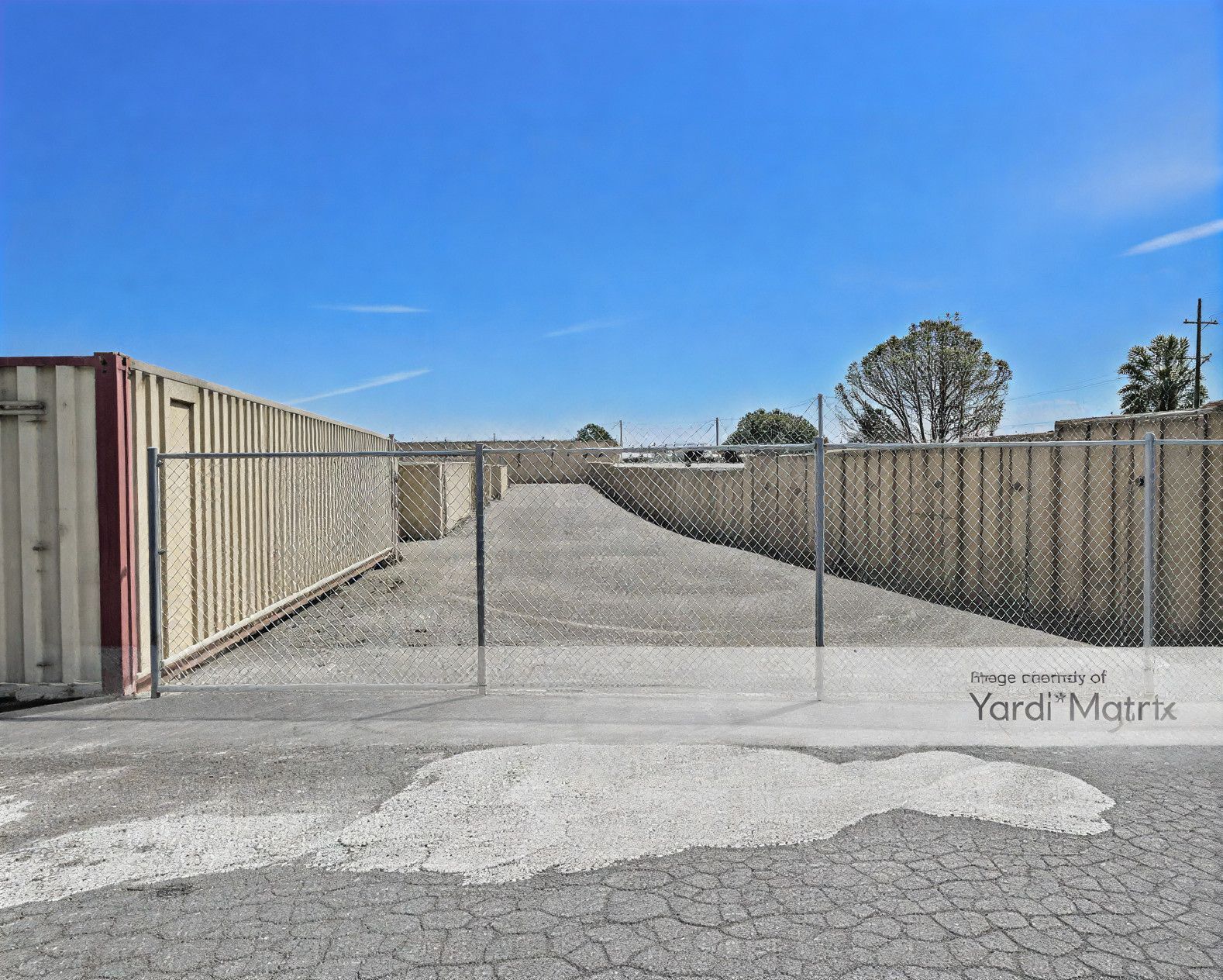 Storage units behind a chain-link fence on a gravel lot under a clear blue sky.