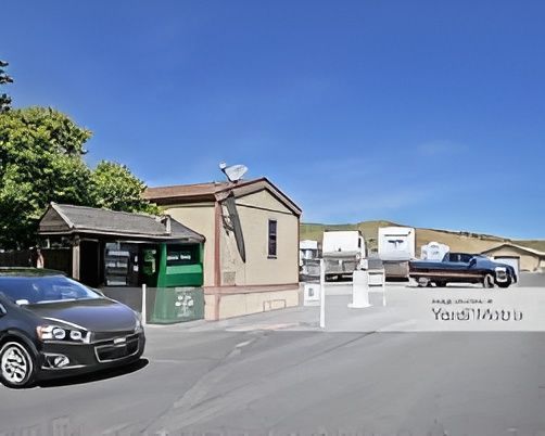 A car parked in front of a building with trailers parked on the right. Blue sky and greenery are in the background.