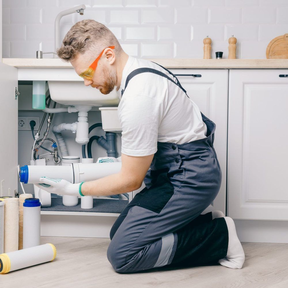 Plumber installing a water filter under a kitchen sink. Wearing safety glasses and work coveralls.