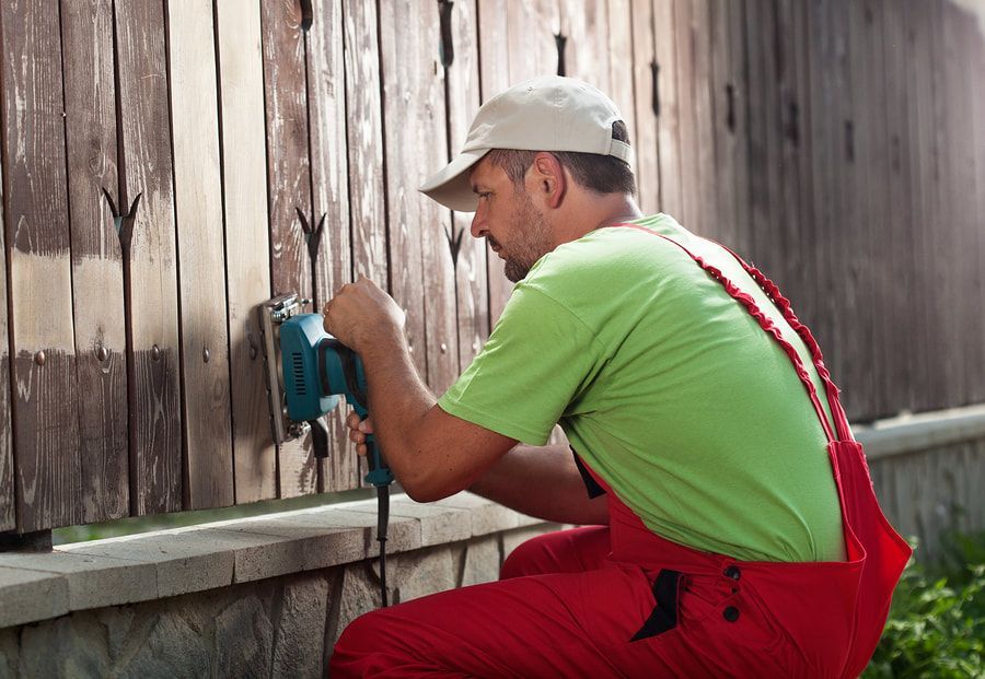 Vancouver Fencing Installation Contractor repairing and refinishing a wood cedar stained privacy fence. The contractor is wearing a green shirt and is sanding the fence for refinishing.