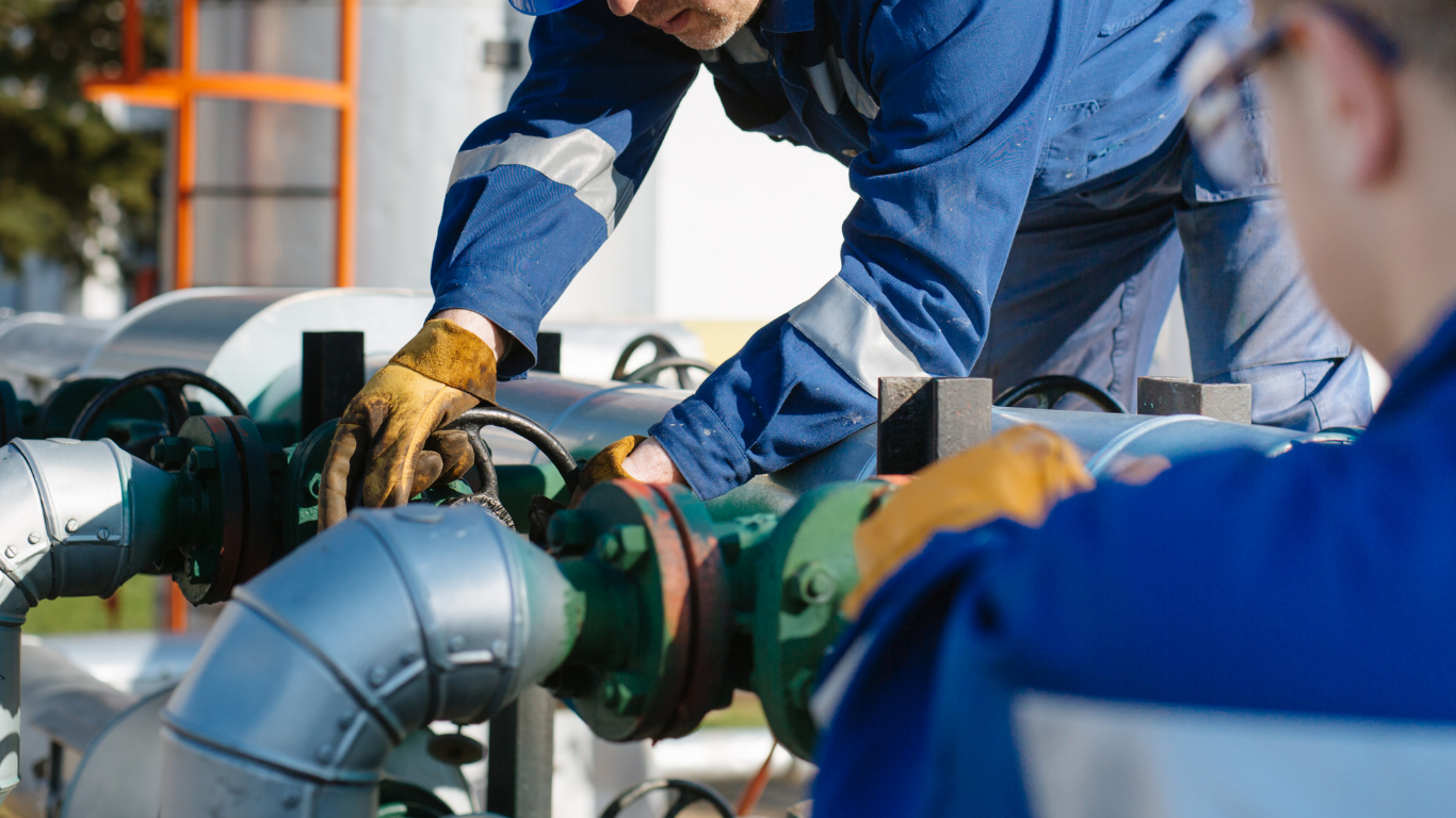 Worker in white protective gear inspecting a large industrial pipeline at a refinery.