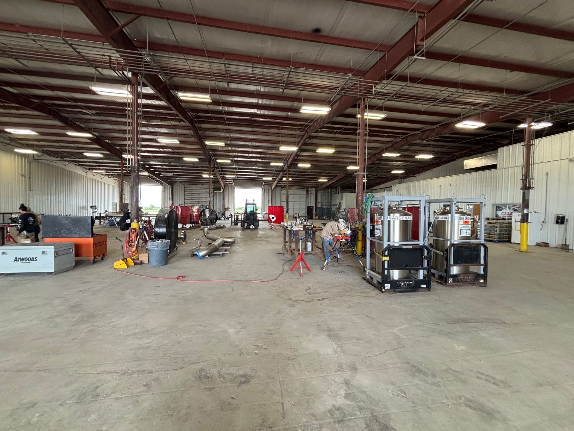 Large industrial warehouse with equipment, work tables, and a few people inside under red steel beams.