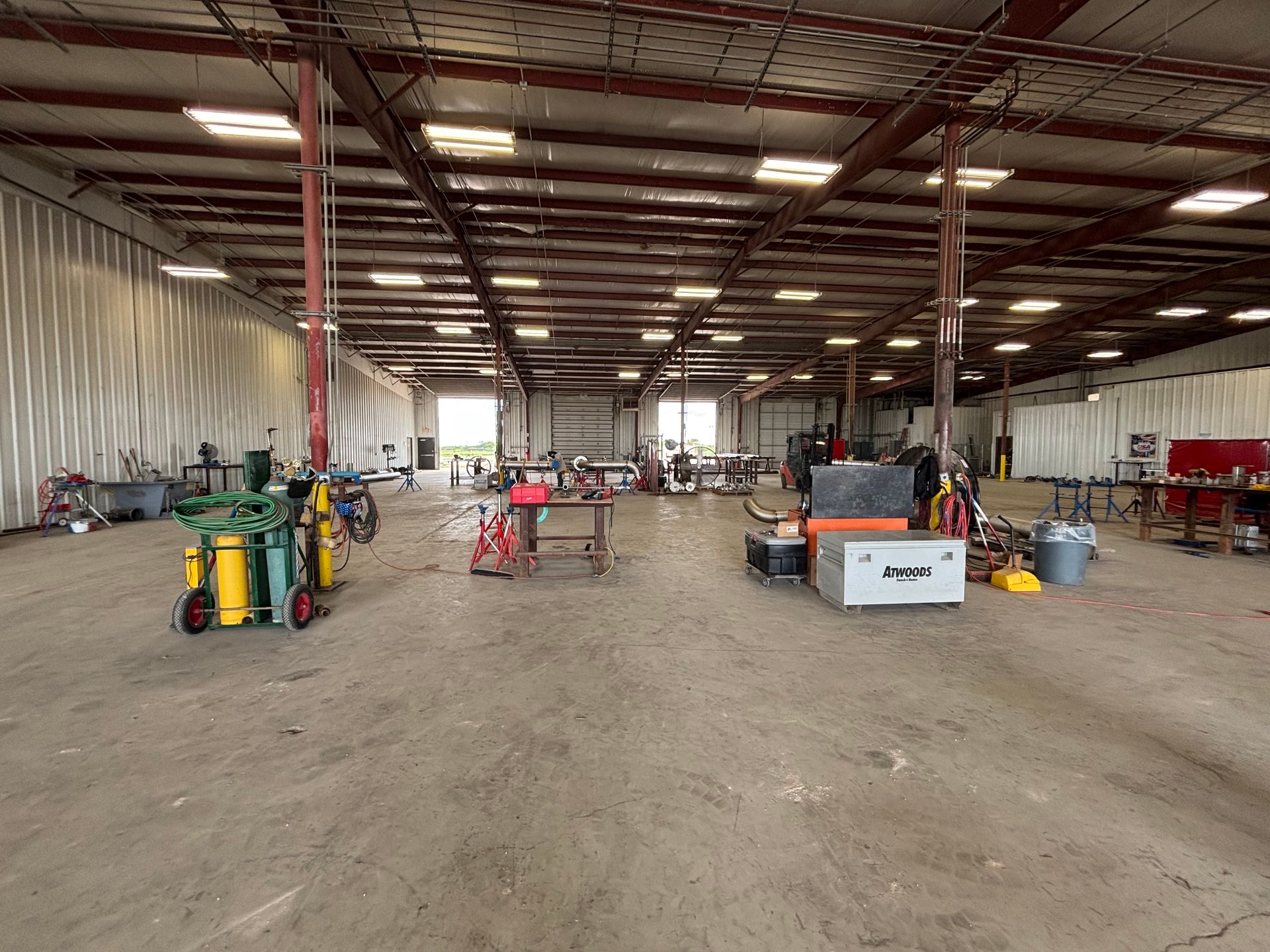 Large industrial warehouse with equipment and tools scattered on a dirt floor under a metal roof.