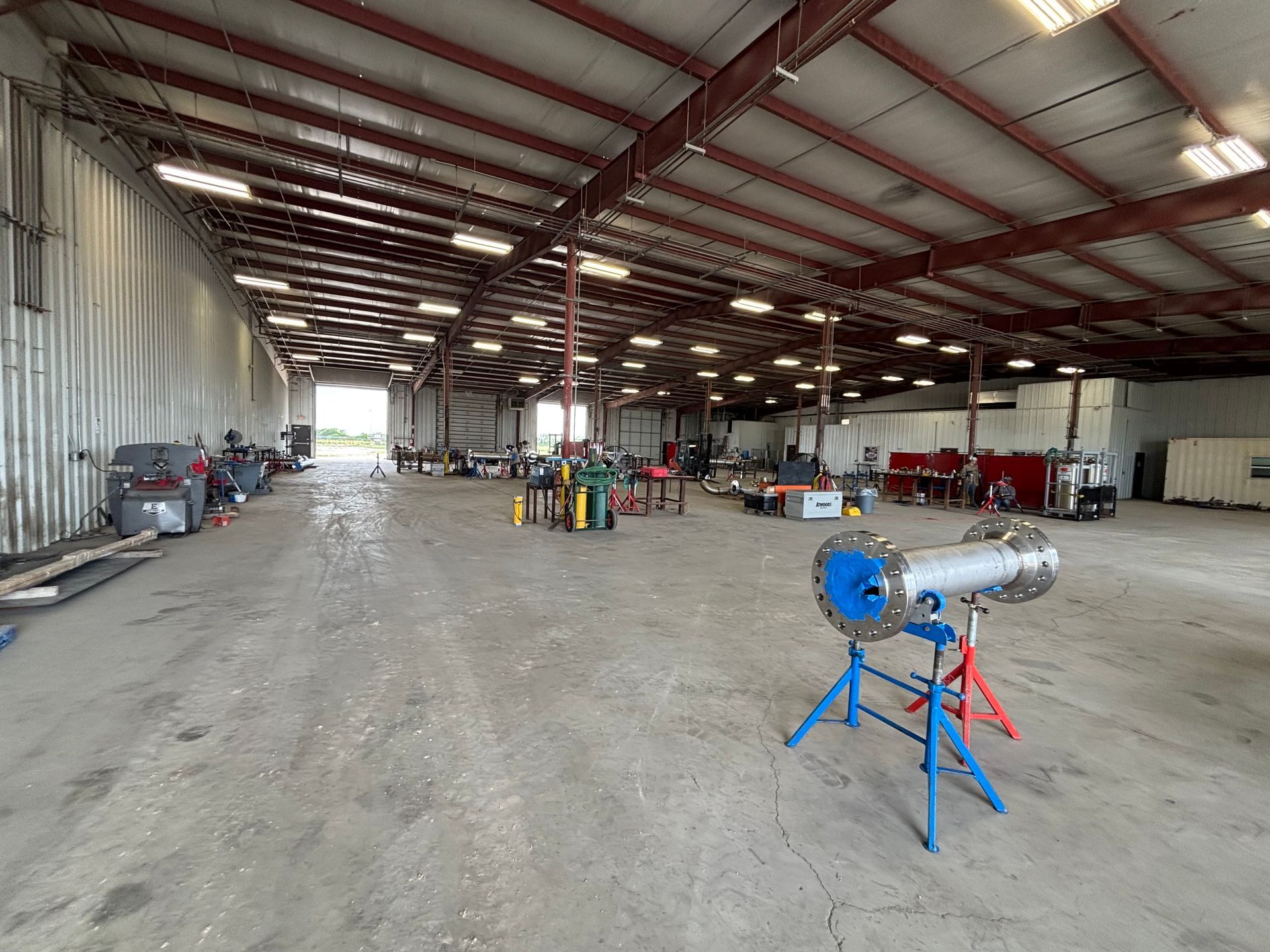Large empty warehouse with metal roof, worktables, and a blue machine on a red stand in the foreground