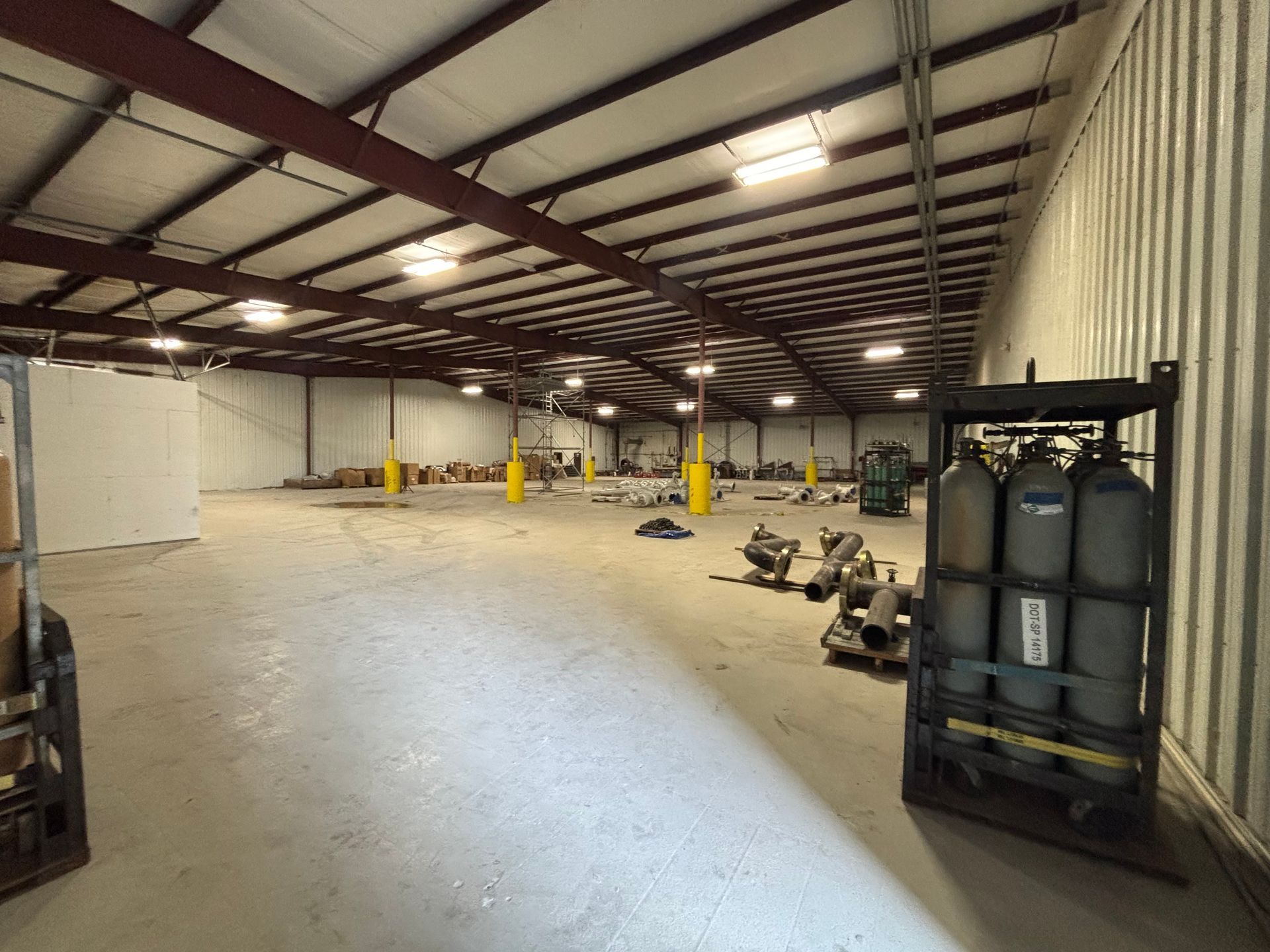 Large empty warehouse interior with concrete floor, metal beams, yellow posts, and equipment near the walls