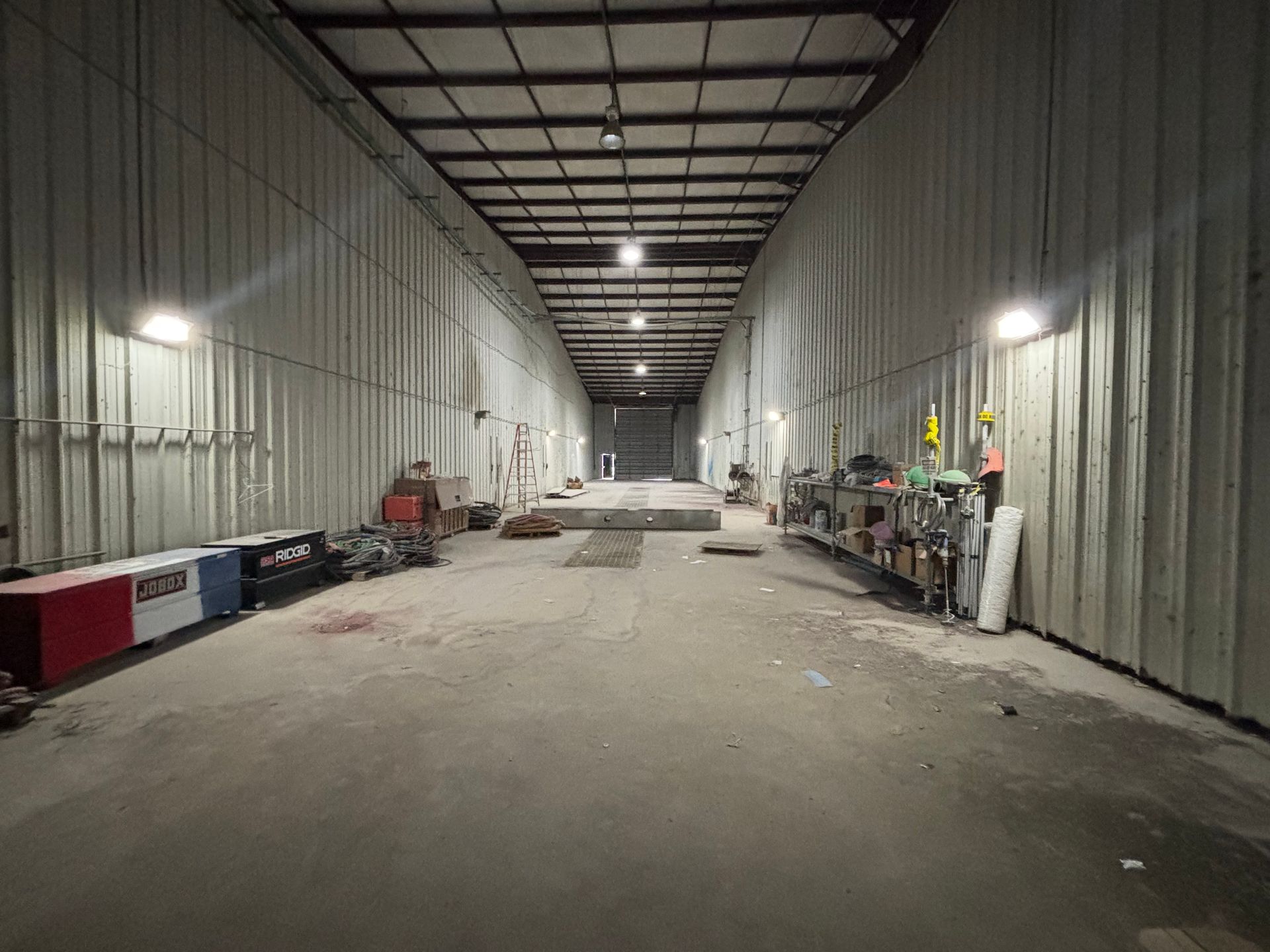 Empty industrial warehouse with high metal walls, concrete floor, and scattered equipment along both sides.