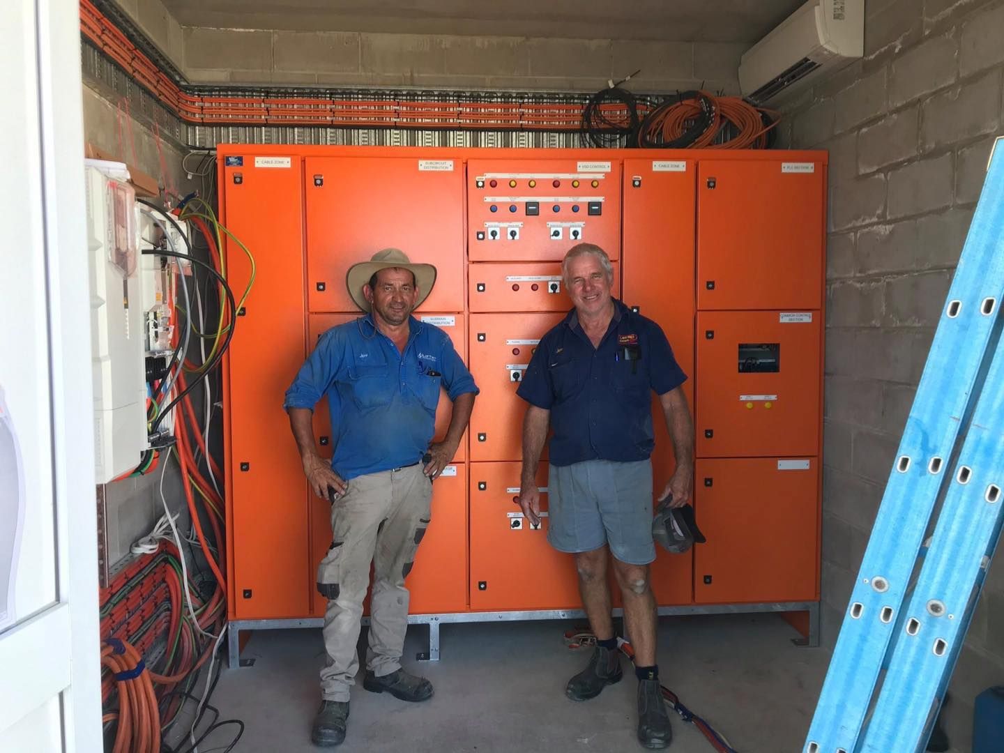 Two Men Stand in Front of an Orange Electrical Panel in a Utility Room — Lahtinen Electrical & Refrigeration in Ingham, QLD
