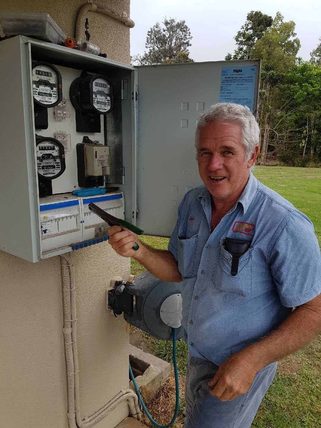 Man in Blue Shirt Points to Electrical Meters in an Outdoor Cabinet — Lahtinen Electrical & Refrigeration in Ingham, QLD