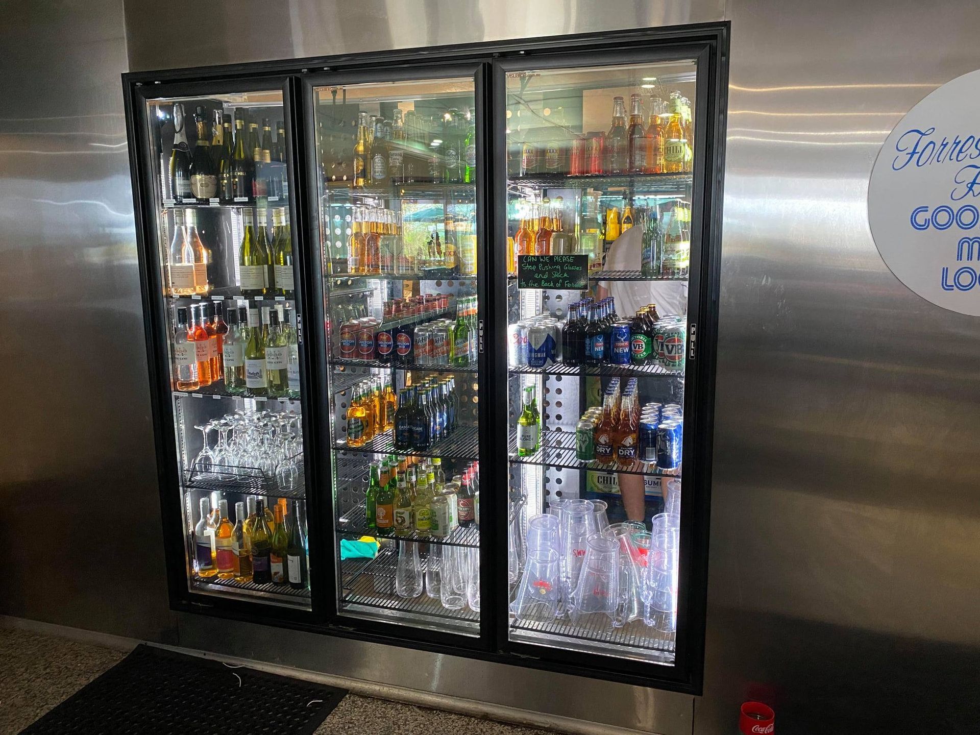 Triple-door glass-front cooler, filled with beverages and glassware, set against stainless steel wall.— Lahtinen Electrical & Refrigeration in Ingham, QLD