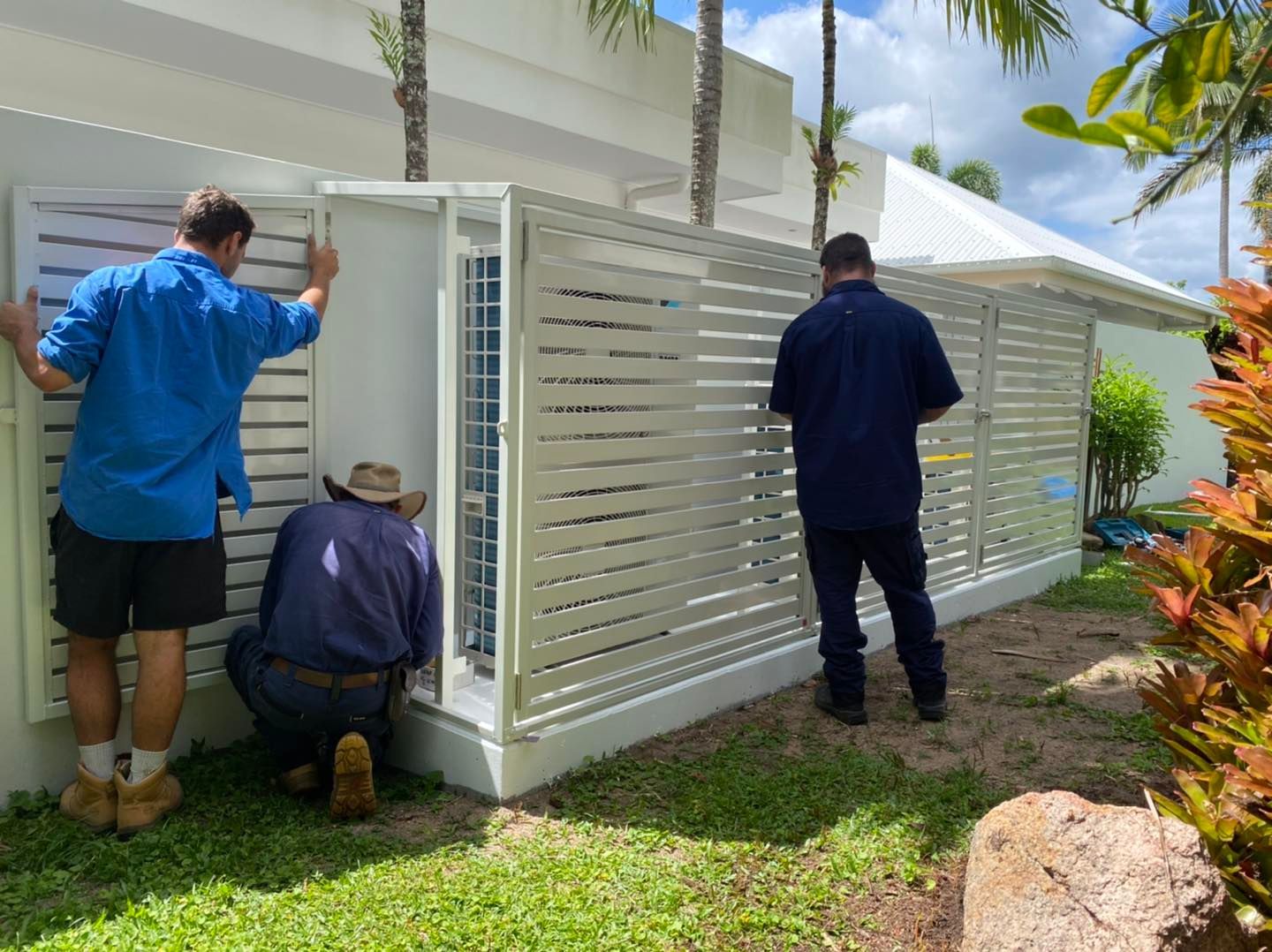 Three People Installing a White Slatted Fence Around an Outdoor Unit — Lahtinen Electrical & Refrigeration in Ingham, QLD