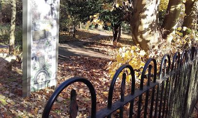 Black iron fence along a wooded park path beside a pale signpost in dappled sunlight