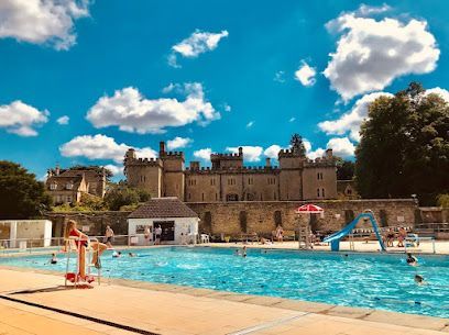 Outdoor swimming pool with swimmers, slides, and a stone castle-like building under a blue sky