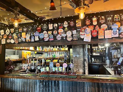 Cozy bar interior with a wooden counter, beer taps, and a wall covered in brewery signs and bottles