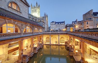 Illuminated historic bath courtyard with a central pool at dusk, stone arches, and warm glowing lights