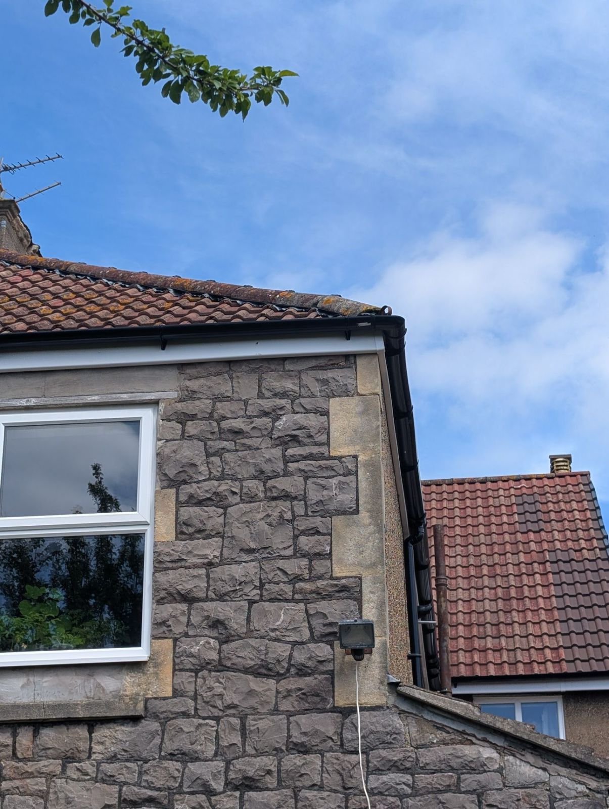 Stone house wall with white-framed window and red-tiled roof under a blue sky
