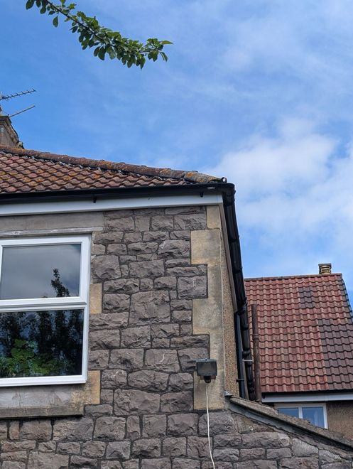 Stone house wall with white-framed window and red-tiled roof under a blue sky