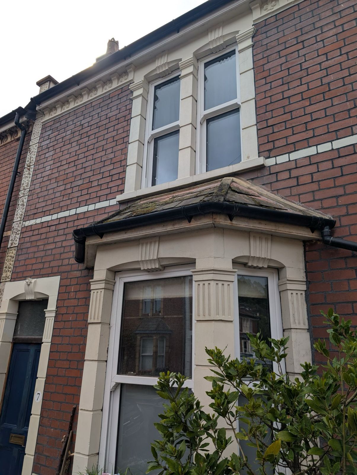 Red-brick house front with a white bay window and small porch roof, viewed from the street.