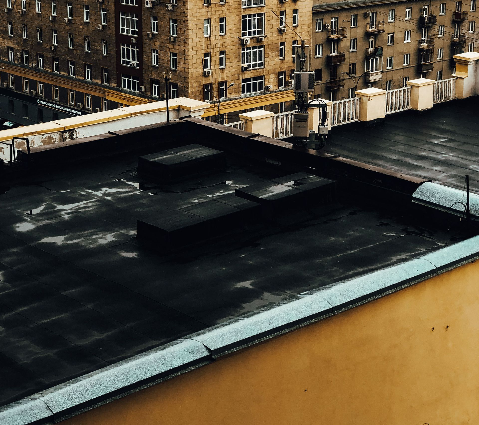 Rooftop with dark vents and a tan parapet, overlooking brick apartment buildings in an urban setting