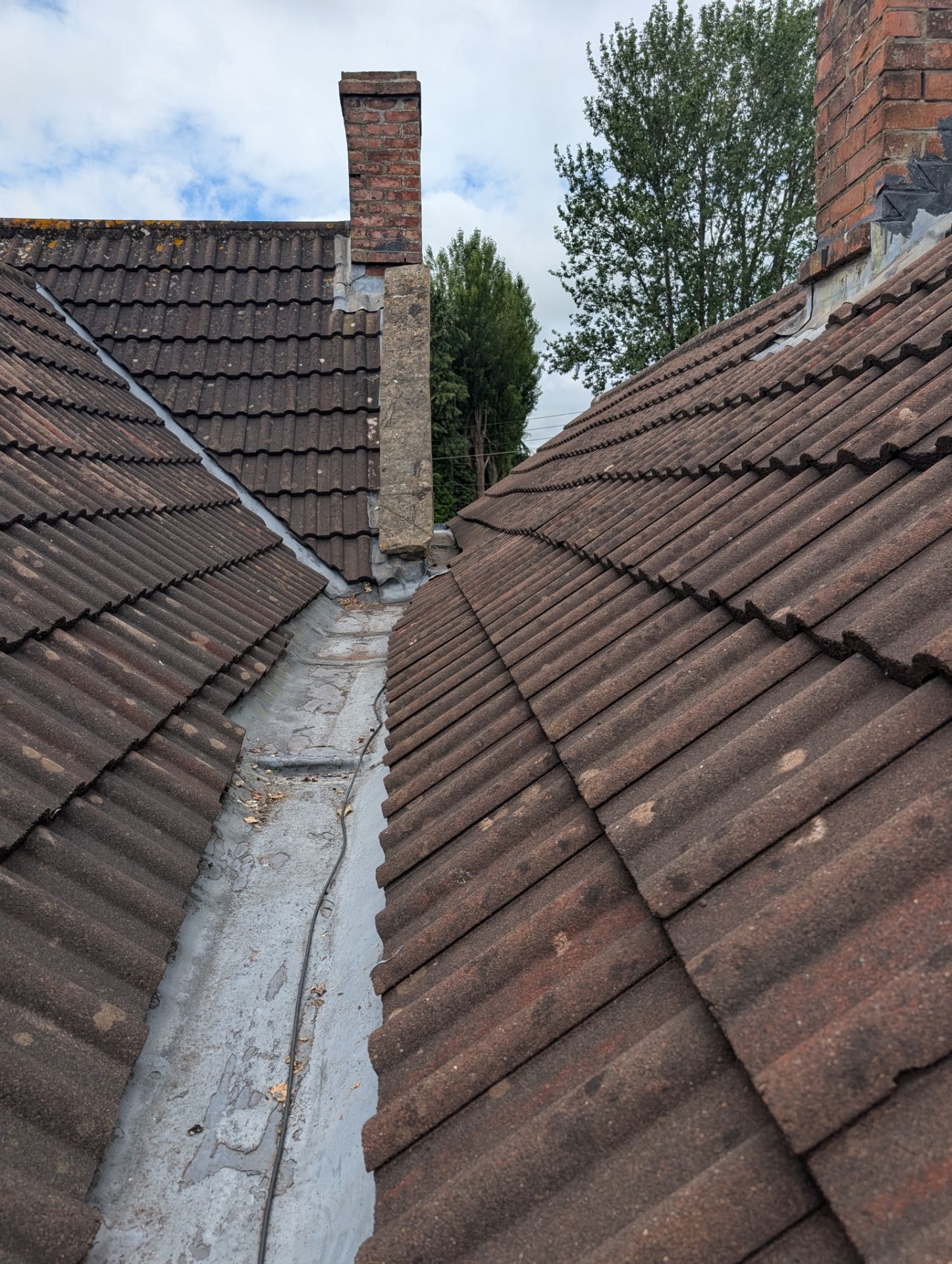 Narrow roof valley between two brown tiled rooftops with a metal gutter and brick chimneys under a cloudy sky