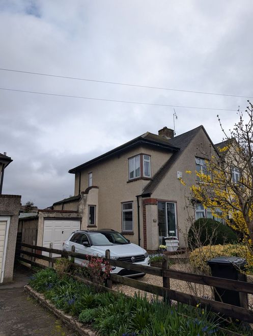 Two-story beige house with a parked silver car and yellow blossoms in a quiet residential street