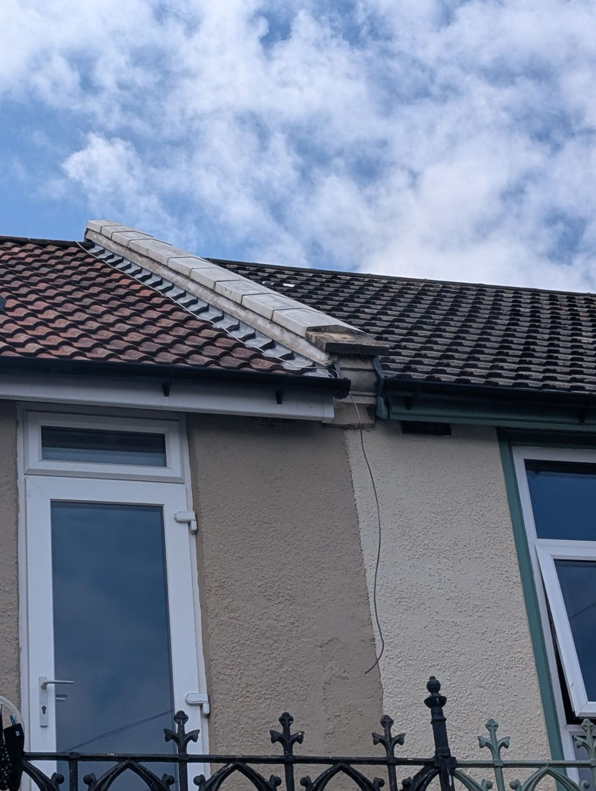 House roof with missing tiles and a damaged ridge near a window, seen above a black iron fence