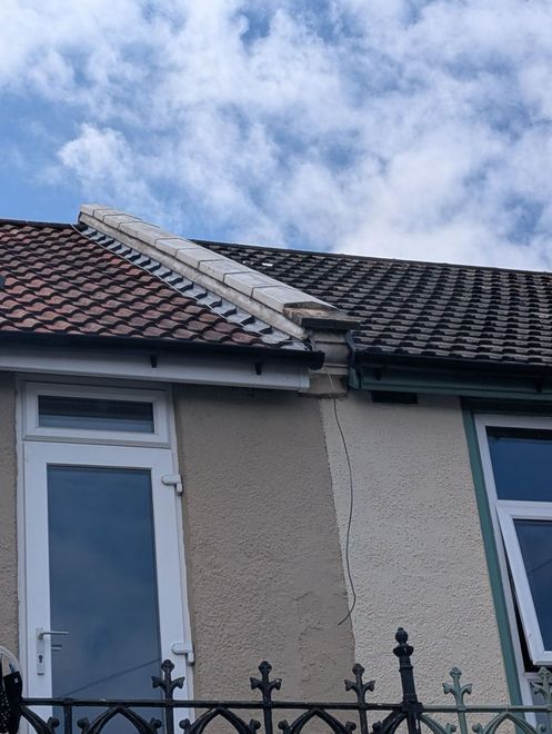 House roof with missing tiles and a damaged ridge near a window, seen above a black iron fence