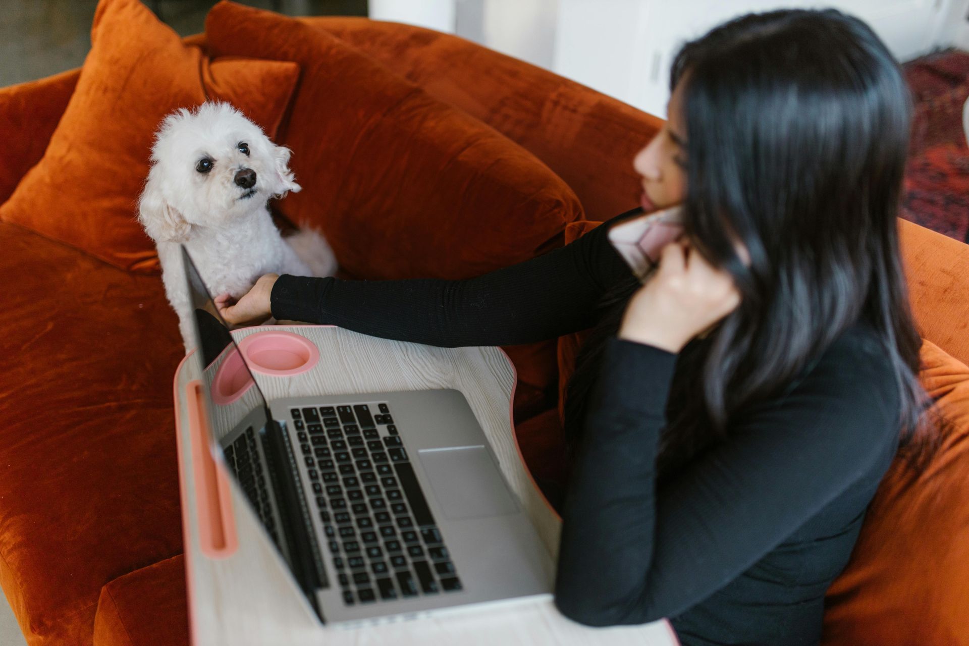 A woman is sitting on an orange couch talking on the phone while holding a laptop next to small white dog.