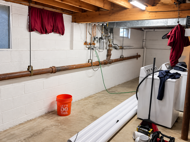 Basement utility room with exposed pipes, orange bucket, white water heater, and stacked PVC pipes.