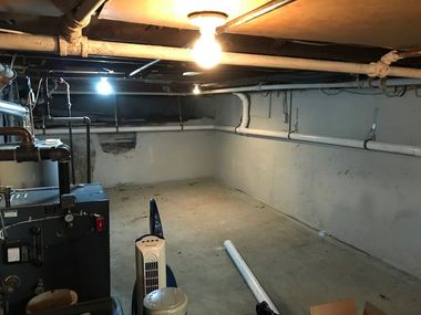 Dim unfinished basement with exposed pipes, a hanging light, and utility items on the floor.