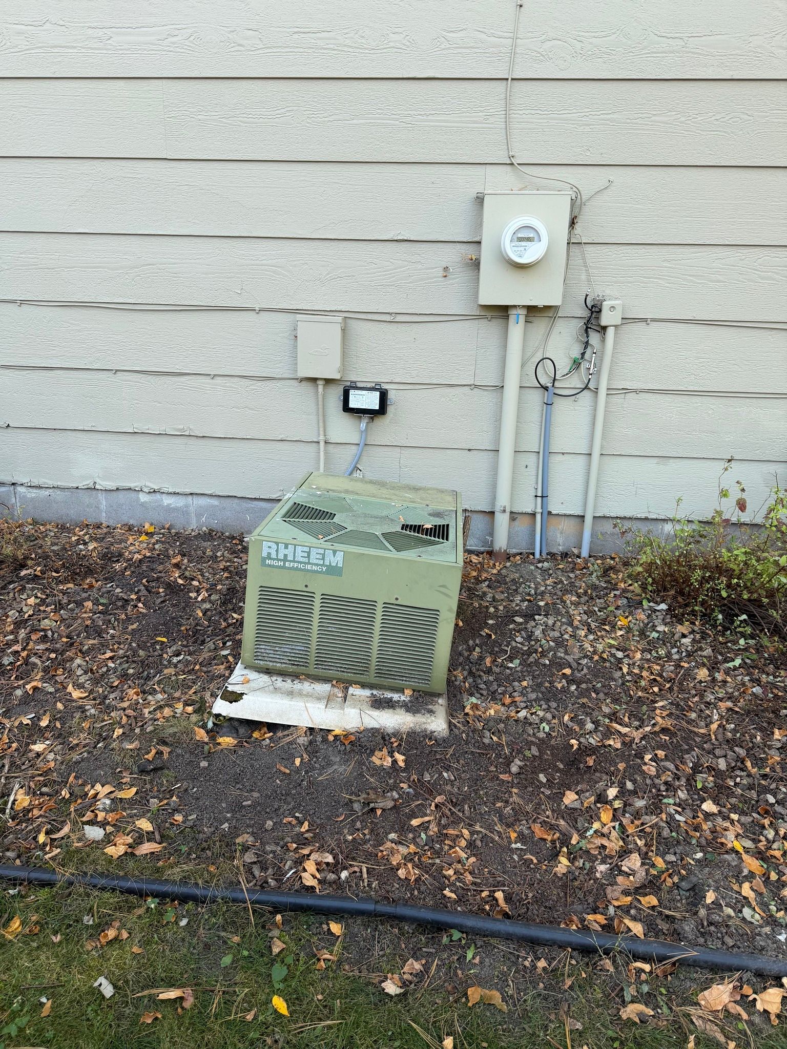 A damaged, green electrical transformer box at base of a building's siding, with other electrical equipment nearby.
