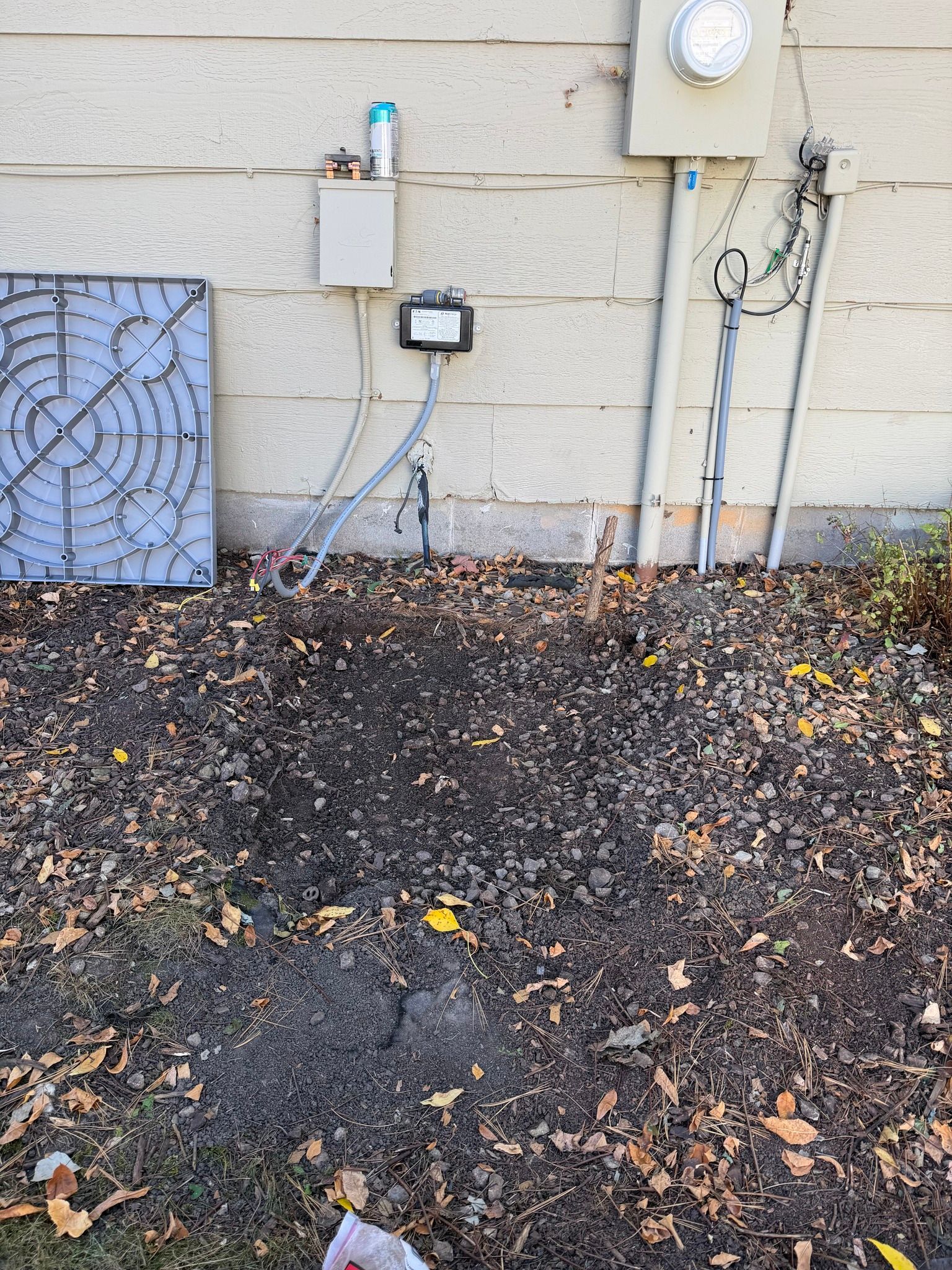 Exterior of a beige building with electrical components and an air conditioning unit. Dirt and fallen leaves cover the ground.