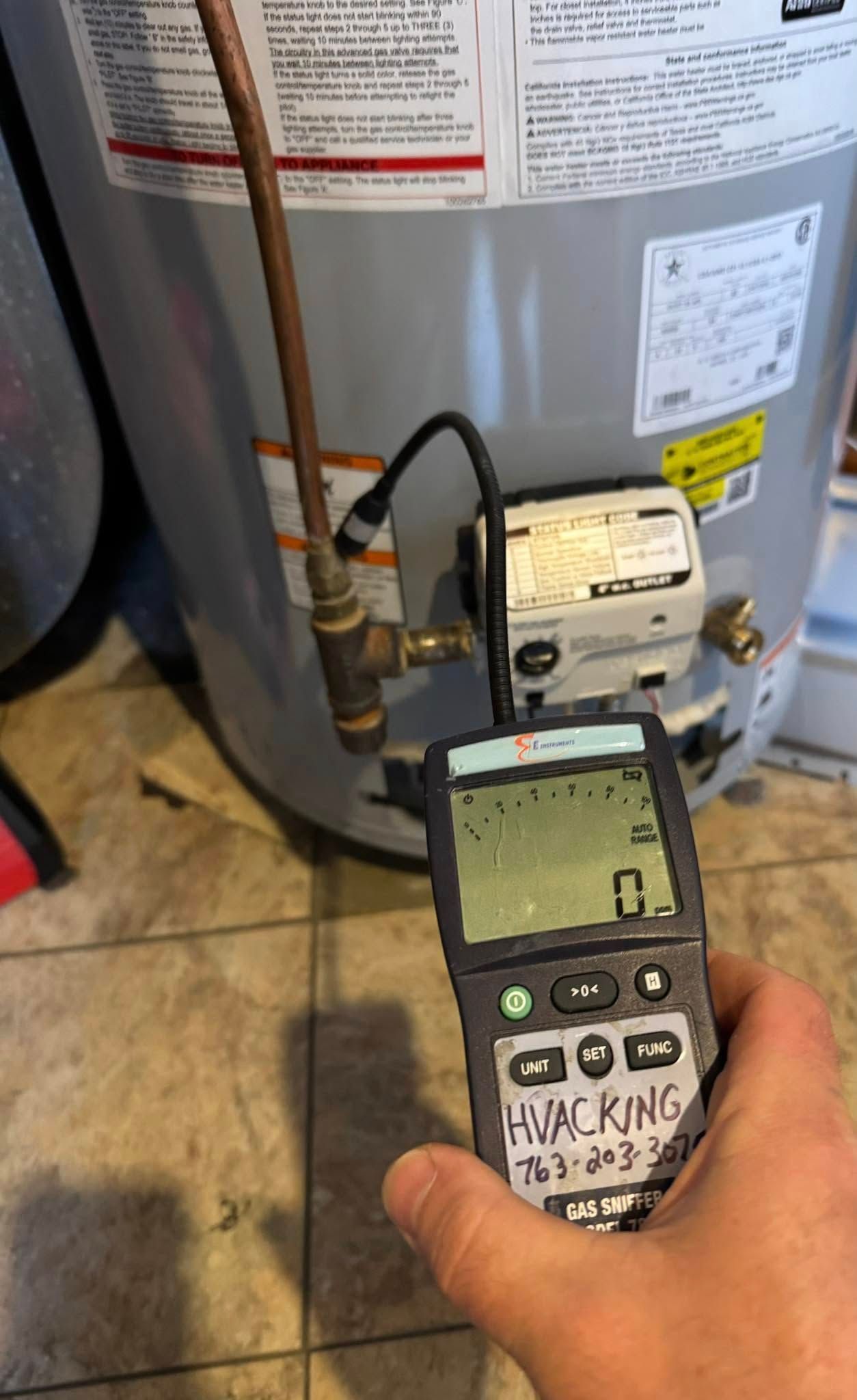 Hand holding a device next to a water heater. The device displays numbers and the hand is labeled 