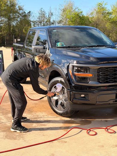 Person washing a black pickup truck with a hose outdoors on a sunny day