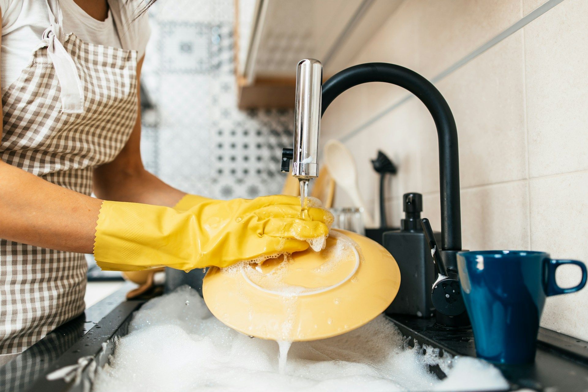 Person wearing yellow gloves washes a yellow plate in a kitchen sink filled with soapy water.