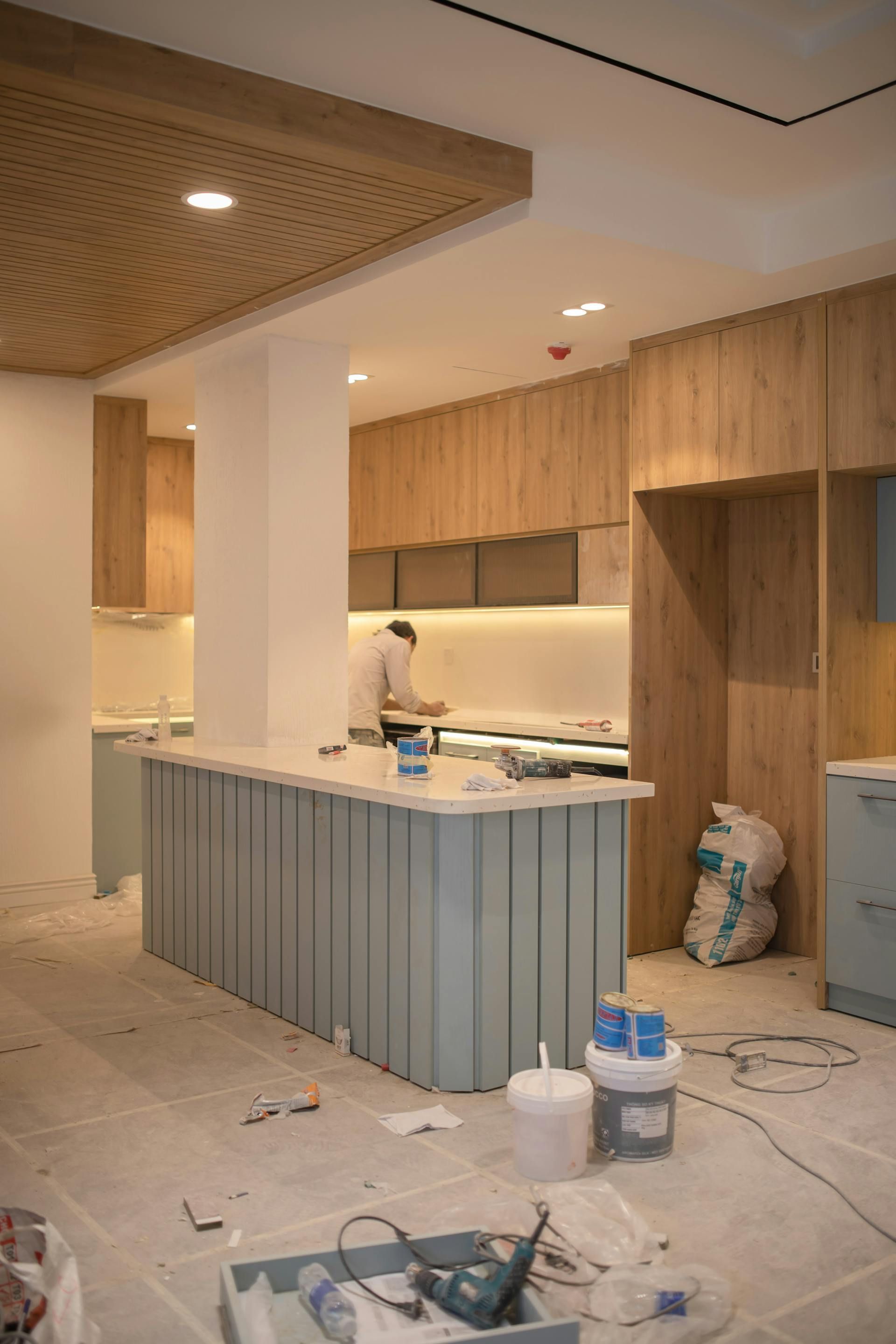 A kitchen under construction; blue island, wood cabinets, worker at counter.