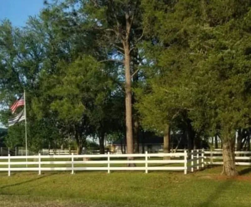 A white fence surrounds a grassy field with trees and a flag in the background.