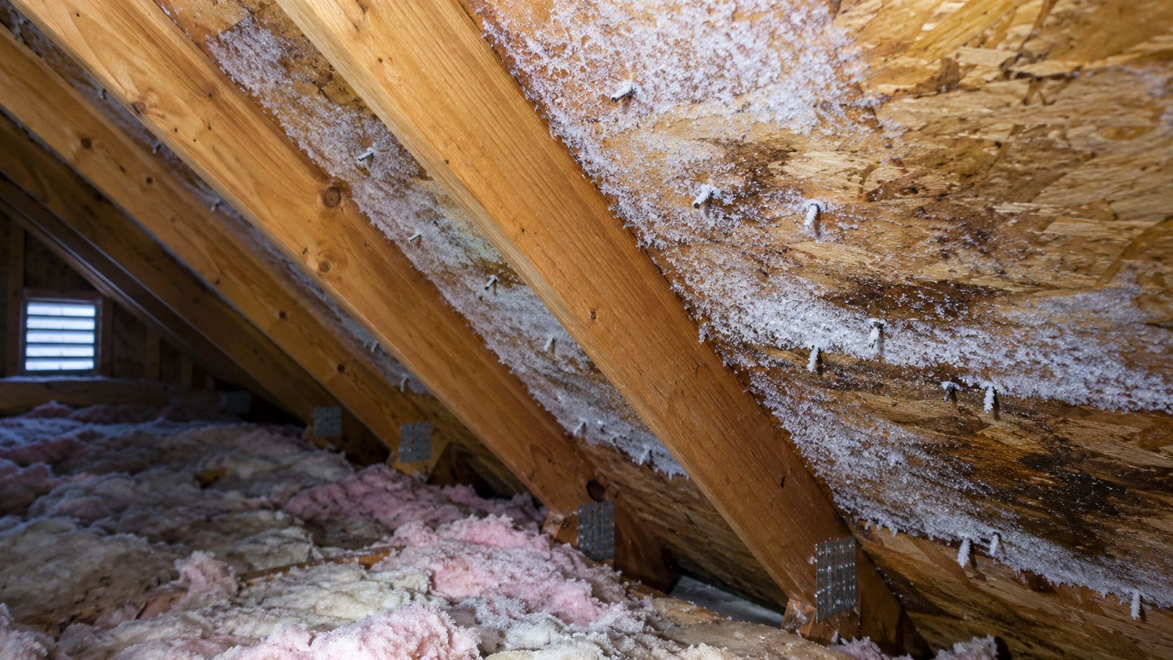 Insulated attic with exposed wooden beams and a small vent window, showing fiberglass insulation on the floor and roof.