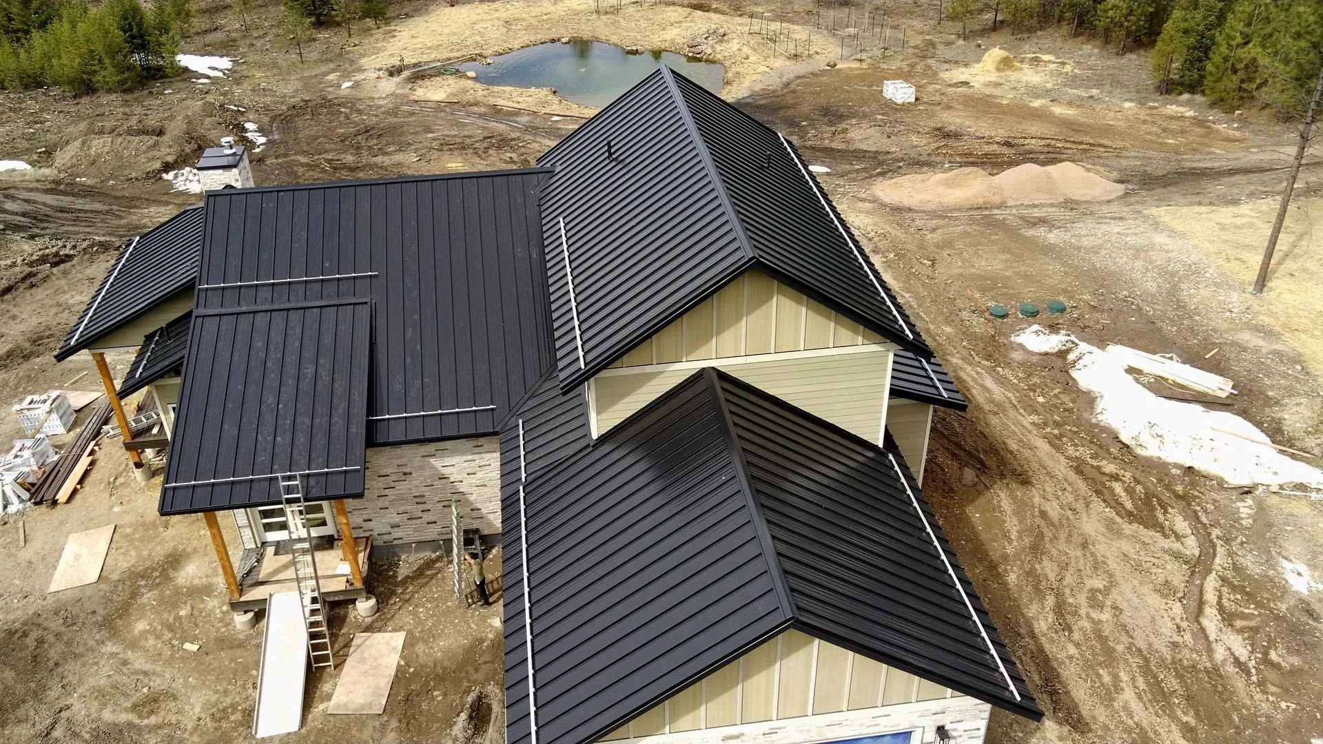 Aerial view of a partially completed house with black metal roofing and exposed framing in a dirt construction site.