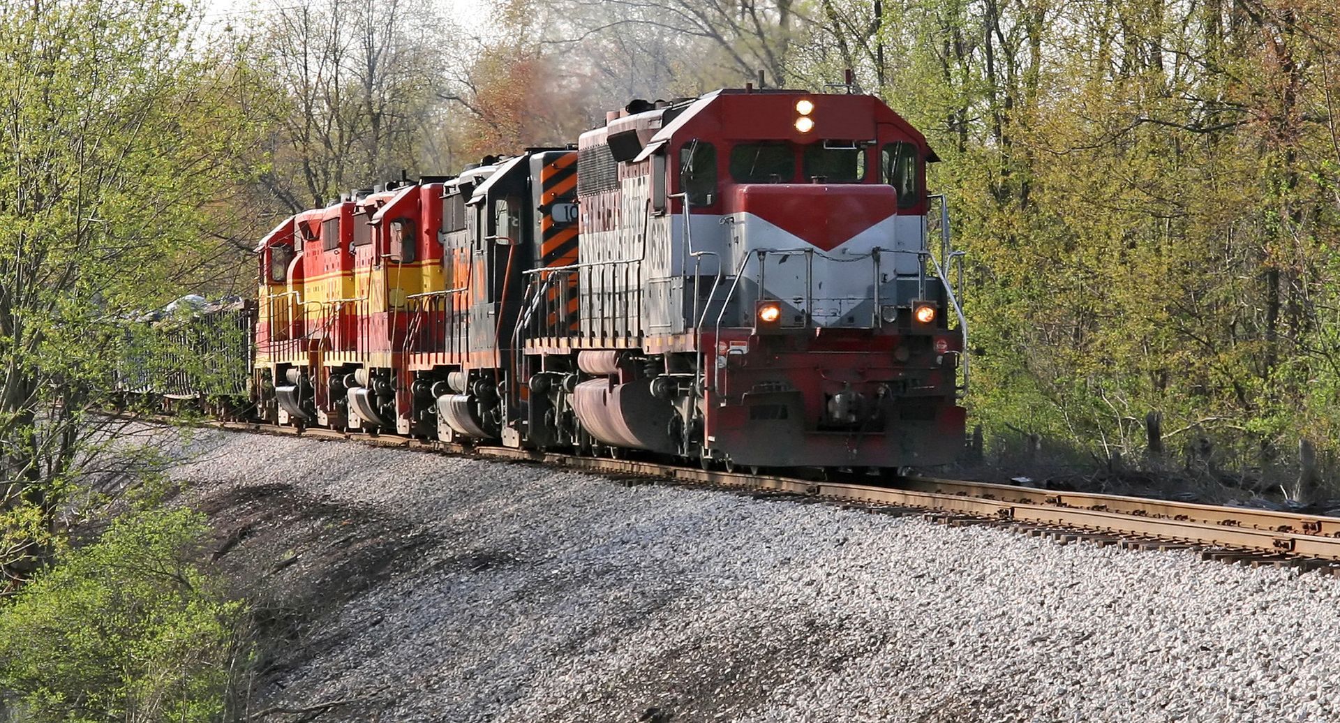 Red and white freight locomotives pulling a train around a gravel track beside trees