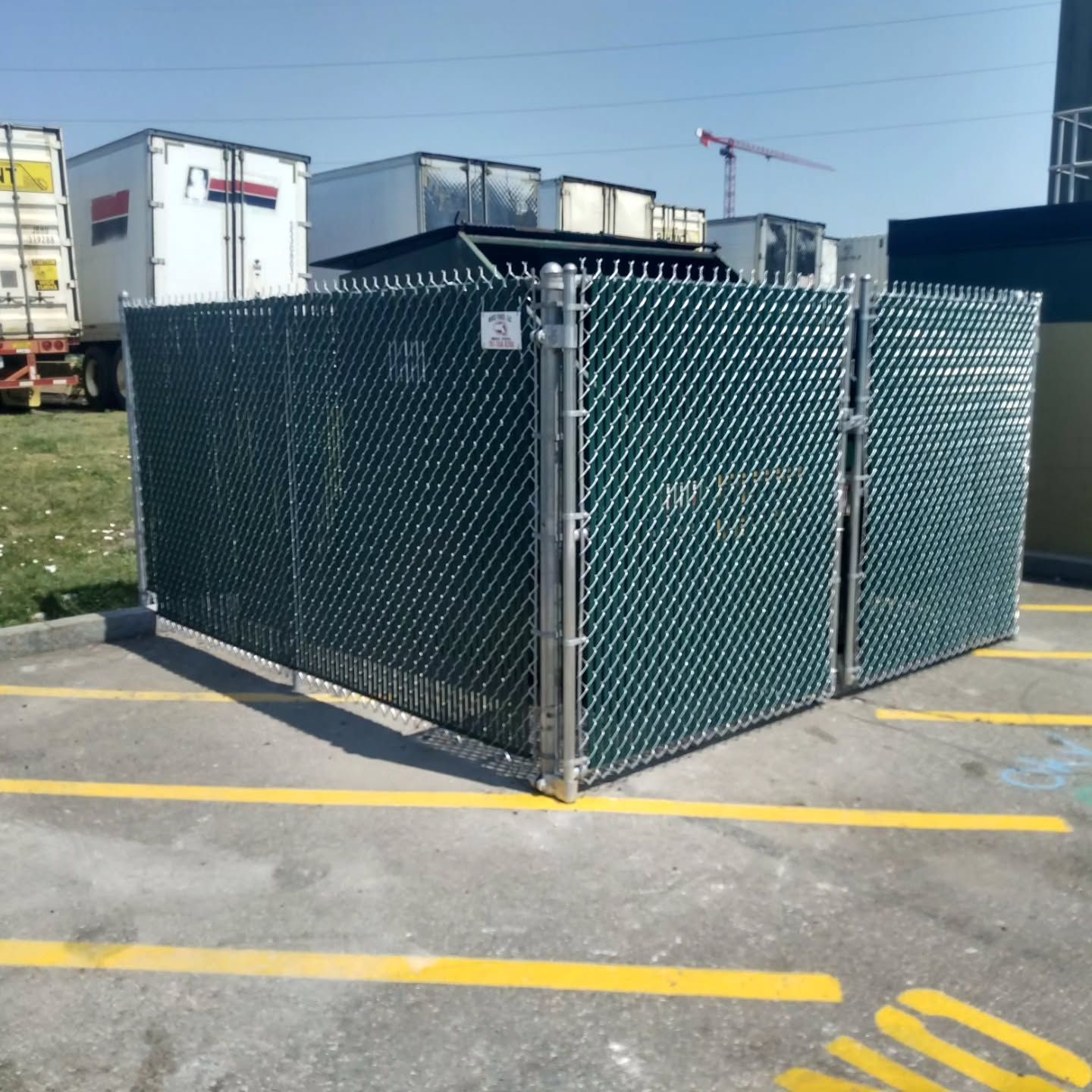Green chain-link fence enclosure in a parking lot with trucks in the background.